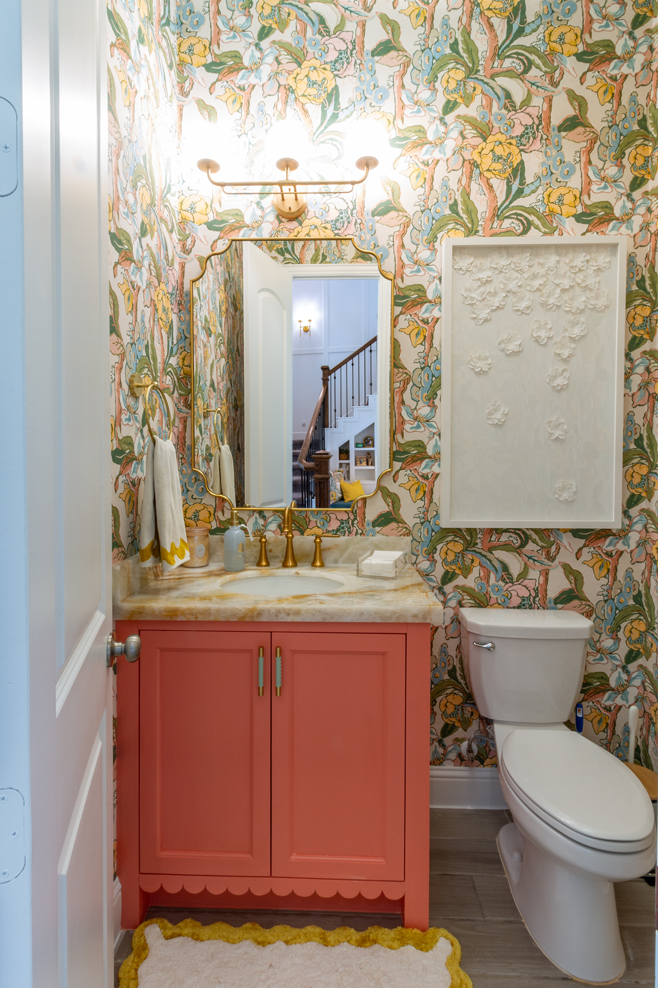 Powder room with botanical wallpaper, a coral-painted vanity, marble top and a scalloped mirror